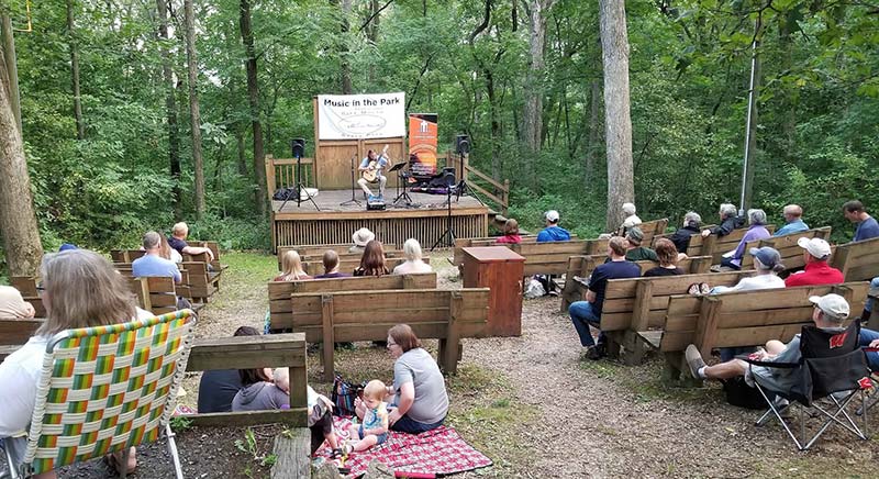 A solo musician plays to a crowd in a small amphitheater in the woods